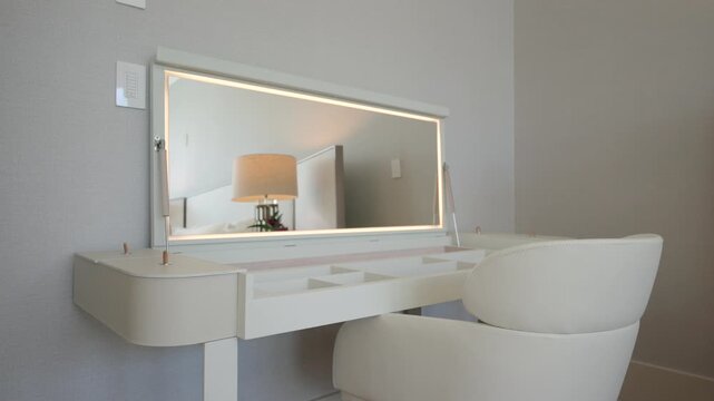 Vanity corner with white table and large mirror featuring LED lighting around edges. Mirror is slightly tilted and reflects part of the room. White cushioned swivel chair