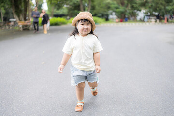 A cute little Asian girl wearing a straw hat walks happily on a walkway in a park.