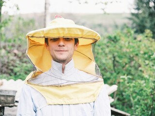A man is wearing a yellow beekeeping suit with mesh and standing outside near beehives. He is...