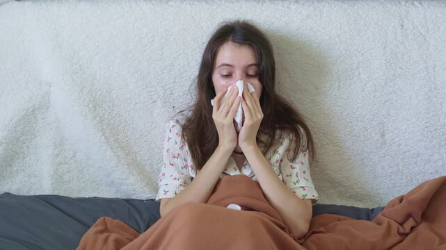 Sick woman blowing her nose with tissue while sitting in bed