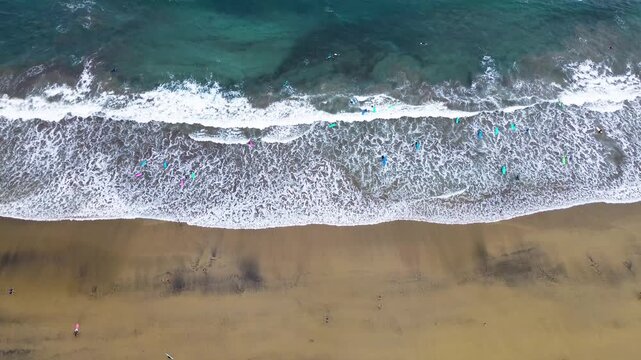 overhead aerial (top-down) view of the active surfing area at a sandy beach (La C&iacute;cer, Las Canteras). The composition is horizontally divided into the dark wet sand, the foamy white line of the waves