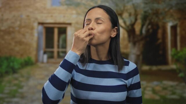 Young hispanic woman outdoors blowing a kiss wearing striped shirt in a rustic outdoor setting with a smile among nature and stone pathway