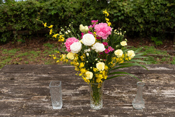 Central crystal vase with white, pink, yellow flowers, flanked by two smaller empty vases. Rustic wood table with green foliage background. Elevated front view, natural daylight. Evokes elegance and h
