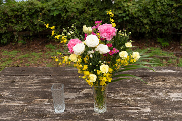Elegant composition: Central crystal vase with white, pink, yellow flowers, smaller empty vase left, on rustic wood. Green foliage background. Elevated front view, natural daylight. Ample copy space r