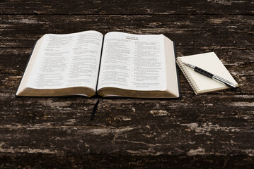 Top view of open Holy Bible (Psalms 149-150, Proverbs 1), notepad with metal quill pen on rustic wood. Natural daylight. Symbolizes spiritual study, wisdom, reflection, devotion.