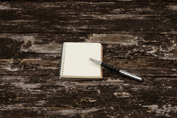 Central notepad with metal quill pen on rustic wood table. Top view in natural daylight. Ample copy space for design. Symbolizes writing, planning, and creative ideas.