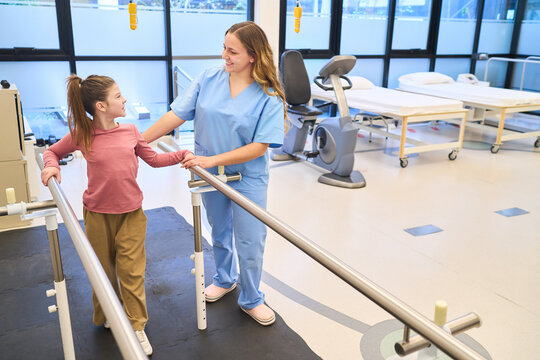 Pediatric physiotherapy session, therapist assisting girl with rehabilitation exercises using parallel bars in a bright clinic, balance training, motor skills development