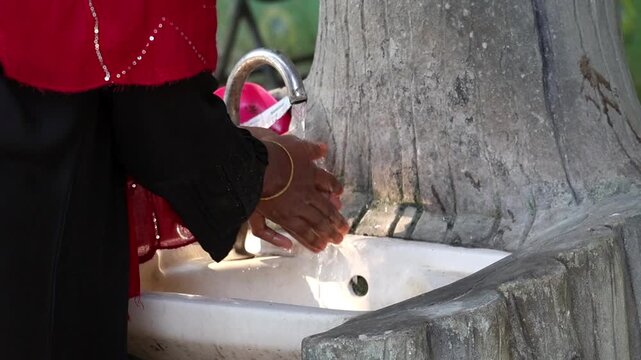 Close-up of an Islamic woman in a traditional black Abaya washing her hands under an outdoor water tap. Emphasizes self-care, religious modesty, purity, and health protection.