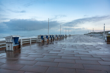 Obraz premium Seaside promenade after rain. Empty promenade with wet tiles and lined beach chairs facing the sea. Calm atmosphere under a brightening cloudy sky.