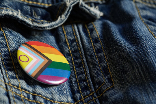 Close-up image of an LGBTQ+ pride badge attached to a blue denim jacket. The pin features the inclusive Progress Pride Flag, representing support, diversity, and the LGBTQ+ community.