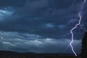 Lightning bolt discharging  towards urban are grounds near tall tree during thunderstorm at night....