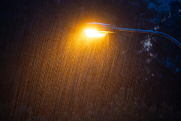 Close-up shot of a street lamp illuminating falling raindrops during heavy rain at night. Bright orange light, dark, moody atmosphere, no people, elevated point of view, long exposure