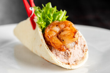 A single, plump grilled shrimp and fresh green lettuce rest on a creamy filling inside a soft tortilla wrap, held together with a red clip on a white plate