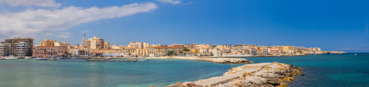 Panoramic Ortigia waterfront in Syracuse, Sicily&mdash;pastel historic buildings, marina and boats behind a stone breakwater under a clear Mediterranean sky; serene coastal cityscape.