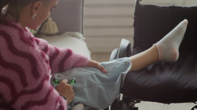Side view shot of young Caucasian girl sitting on couch in living room and using triangle ball massager applying gentle pressure on legs placed on wheelchair nearby for better recovery