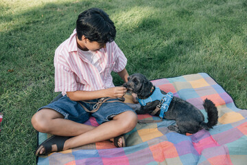 Teenager feeding pet dog with treat while sitting on a colorful blanket in a park