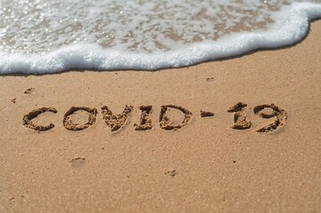 Fototapeta premium Close-up of the phrase covid 19 written in sand on a shoreline. Summer travel limitations caused by outbreaks of respiratory illnesses. Isolation measures in warm regions. Immunization campaigns
