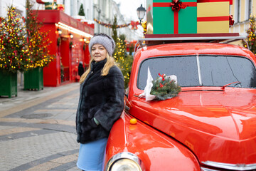 A white woman stands next to a vintage red car in a festive urban setting decorated for the...