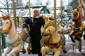 A middle-aged white woman stands next to colorful merry-go-round horses against a snowy landscape. The scene conveys a festive winter atmosphere. © Светлана Высокос