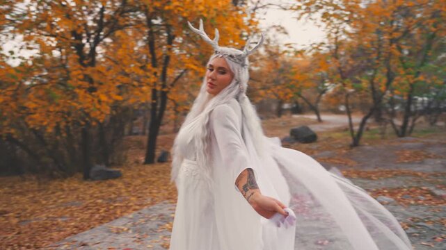 fantasy young elf woman beauty face walking in autumn nature yellow trees foliage. sexy adult fairy tale girl long blonde ash hair white dress snow queen style makeup. myth faun deer antlers on head