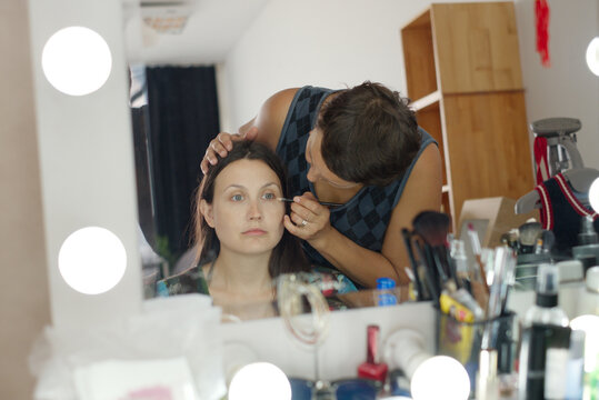 Caucasian young adult woman sitting while Caucasian young adult man applying makeup to her face in front of mirror, various cosmetic products and tools visible on table