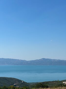 A serene panoramic view of a turquoise lake surrounded by lush green hills and distant blue mountains under a clear summer sky, capturing the calm beauty of untouched nature in perfect harmony.