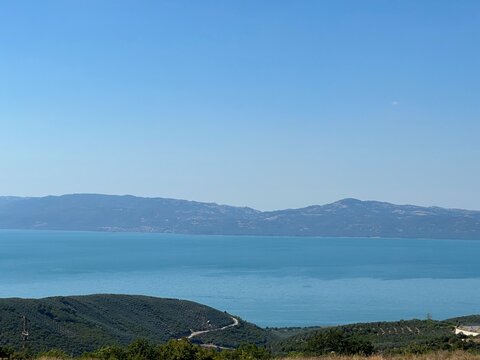 A serene panoramic view of a turquoise lake surrounded by lush green hills and distant blue mountains under a clear summer sky, capturing the calm beauty of untouched nature in perfect harmony.