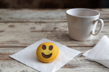 Shortbread cookie shaped like a smiley emoji lying on a white napkin on a wooden table next to a white cup, close-up, selective focus.