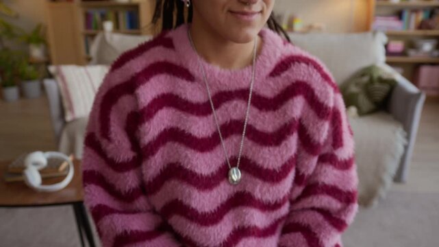 Tilt up portrait of young Caucasian girl with dreadlocks wearing bright cozy sweater smiling while posing for camera in wheelchair in modern living room interior