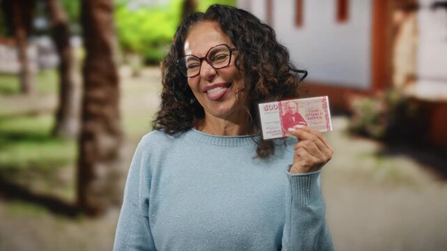 Middle-aged woman playfully showing icelandic krona on a city street, capturing a vibrant moment with a cheerful expression, glasses, and a knit sweater in an outdoor urban background.