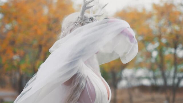 elf woman beauty face walking in autumn nature yellow trees. sexy fairy tale girl long blonde ash hair white silk dress waving cape train, snow queen style makeup. myth faun deer antlers on head