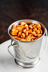 A closeup of spicy roasted peanuts in a small metal serving bucket with a handle, sitting on a white plate against a dark, out-of-focus background.