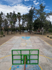 Vertical drone view of an outdoor basketball court in Zapopan, Mexico. Concrete surface, yellow lines, blue key, surrounded by park trees and benches under a cloudy sky