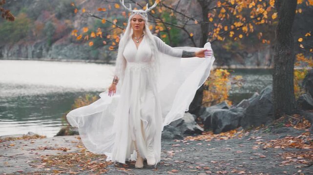 fantasy young elf woman walking in autumn nature yellow trees foliage. sexy adult fairy tale girl long blonde hair, waving cape veil silk white dress snow queen style. myth faun deer antlers on head