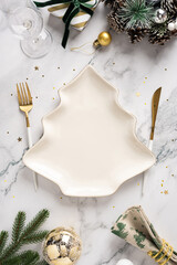 A vertical festive Christmas table scene features a unique tree shaped plate, gold cutlery, ornaments, pine branches, and sparkling decor for New Year celebration and holiday greeting