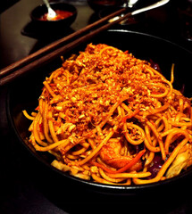 Close-up of flavorful Indian-Chinese style spicy garlic noodles topped with crispy fried garlic and served in a black bowl. The rich colors, texture, and moody lighting create an appetizing food photo