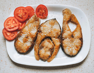 Pieces of sesame-fried fish served on a white plate with sliced tomato. A perfect dish for breakfast, lunch, or dinner.
