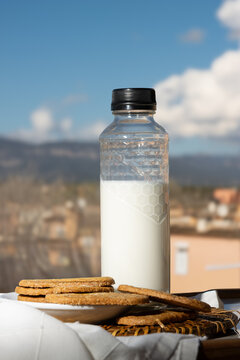 Botella de leche y galletas integrales sobre una mesa al aire libre con un fondo de paisaje mediterr&aacute;neo y cielo azul. 