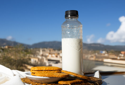 Botella de leche y galletas integrales sobre una mesa al aire libre con un fondo de paisaje mediterr&aacute;neo y cielo azul. 