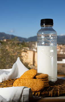 Botella de leche y galletas integrales sobre una mesa al aire libre con un fondo de paisaje mediterr&aacute;neo y cielo azul. Concepto de desayuno saludable, natural y artesanal en un entorno rural soleado.