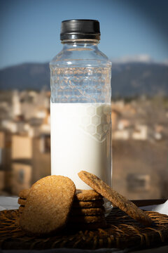 Botella de leche y galletas integrales sobre una mesa al aire libre con un fondo de paisaje mediterr&aacute;neo y cielo azul. 