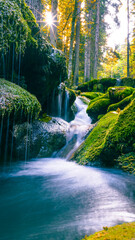 Mossy rocks and waterfall illuminated by sunlight streaming through dense autumn forest