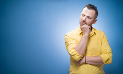 Doubt young man looking to side, thinking with frowned face expression, copy space for text over blue background, dresses in yellow shirt