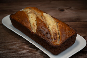 A freshly baked banana bread gateau is displayed on a white plate, showcasing its golden-brown crust and ripe banana slices on top. Perfect for a sweet treat or dessert.