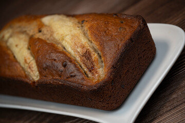 A freshly baked banana bread gateau is displayed on a white plate, showcasing its golden-brown crust and ripe banana slices on top. Perfect for a sweet treat or dessert.