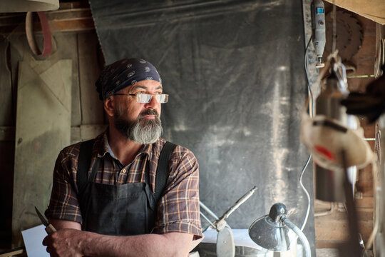 Craftsman with beard in workshop, wearing bandana and apron, holding a knife, looking thoughtful, artisan at work, small business owner, maker, entrepreneur, handmade