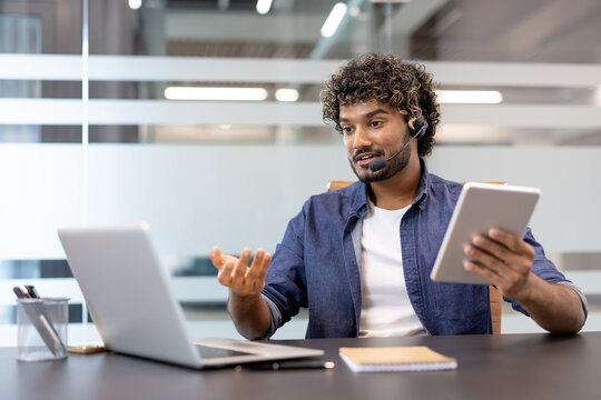 A young Indian man in a suit is sitting in an office, holding a tablet in his hand and talking on a video call on a laptop