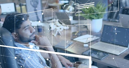 Leaning South Asian man processing network data at modern office desk, with laptop, monitors, plant