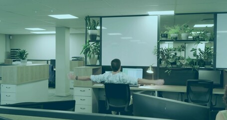 Stretching man wearing shirt and trousers extending arms at open-plan desk, with monitors, plants