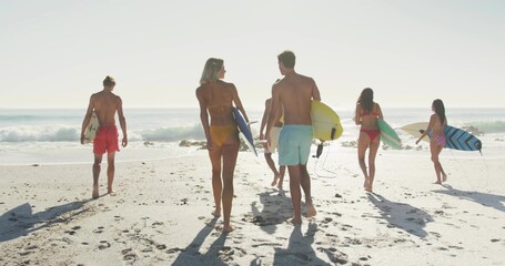 Walking fit group of six surfers carrying colorful surfboards on sandy beach, showing swimwear